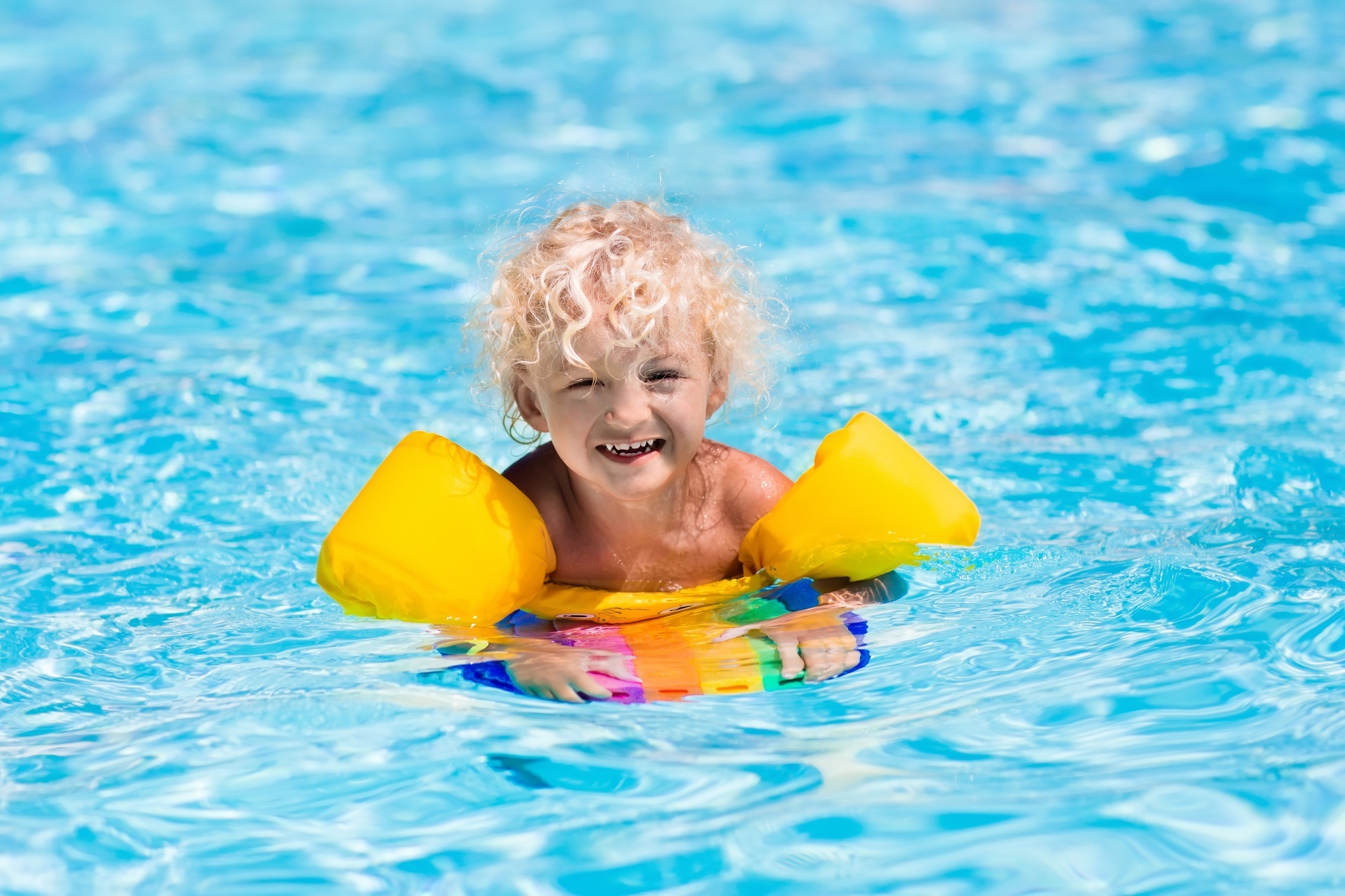 Happy laughing little boy playing in outdoor swimming pool on a hot summer day. Kids learn to swim. Child with colorful floaties and floating kickboard. Family vacation in tropical resort.
